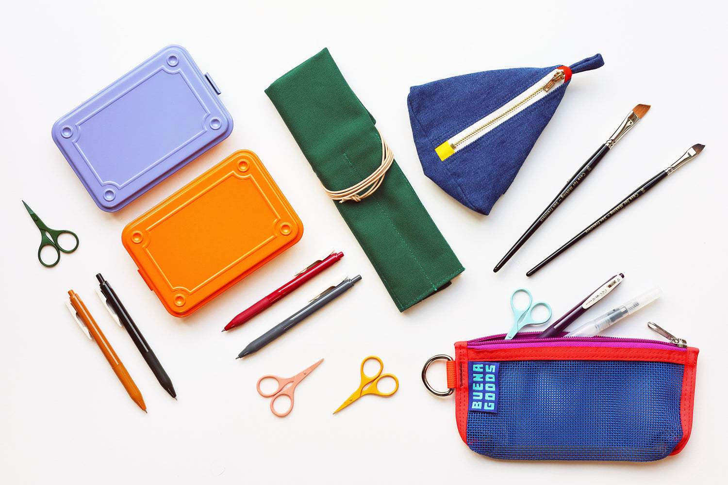 A flat-lay arrangement of colorful stationery and art supplies on a white background, including two colorful metal boxes, a green brushc roll, a blue denim pouch with a pencil, paintbrushes, pens, and a blue mesh pencil case with 'BUENA GOODS' printed on it.