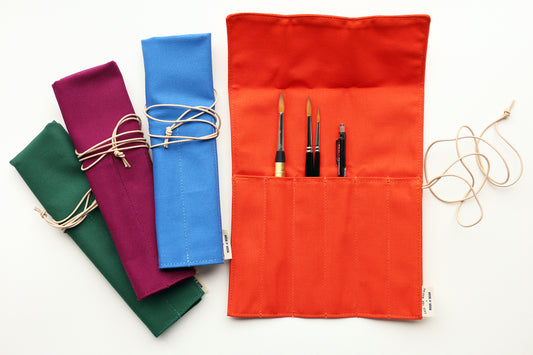 A set of fabric art supply rolls in multiple colors, including orange, blue, purple, and green, shown on a white background. The orange roll is open, displaying pockets holding paintbrushes and a pencil, with a beige  leather cord tied around it.
